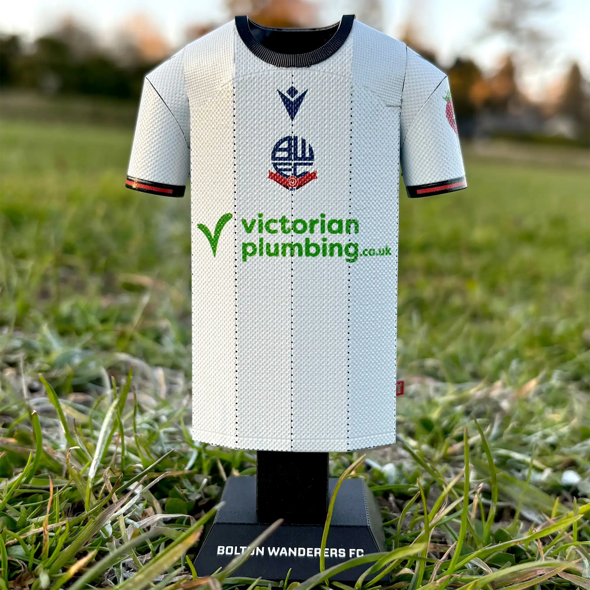 Square lifestyle product photograph of a retro white Bolton Wanderers FC jersey collectible styled in an outdoor grassy setting.