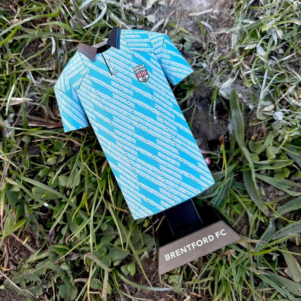 Square lifestyle product photograph of the retro light blue Brentford FC jersey collectible styled in an outdoor setting.