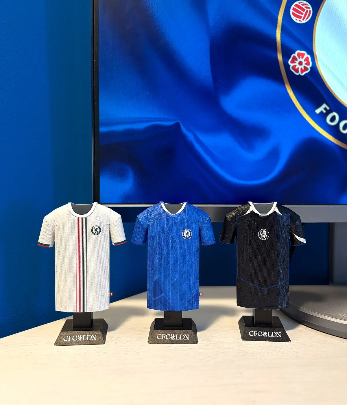 Three miniature football shirts on stands with a large screen displaying a logo in the background.