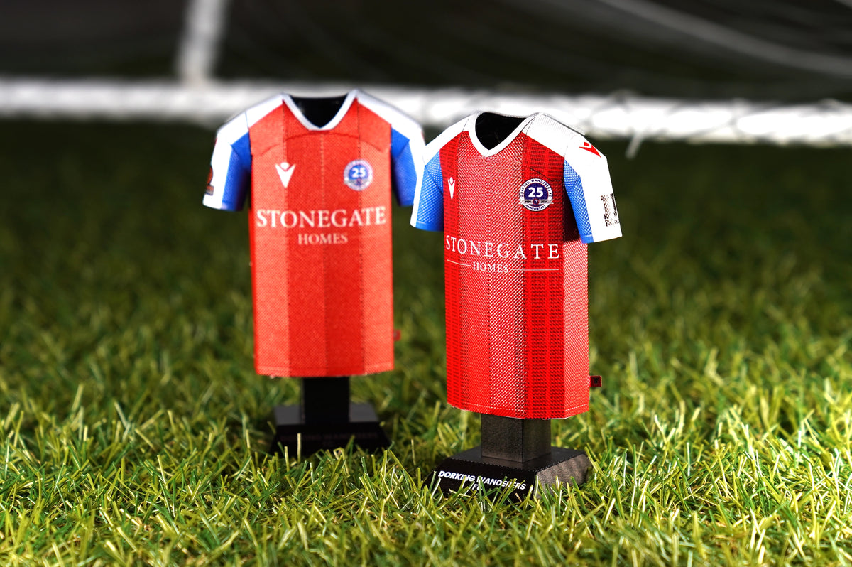 Desktop web lifestyle product photograph showcasing a pair of Dorking Wanderers FC 2024/2025 home jersey collectibles displayed together on an outdoor grass turf.