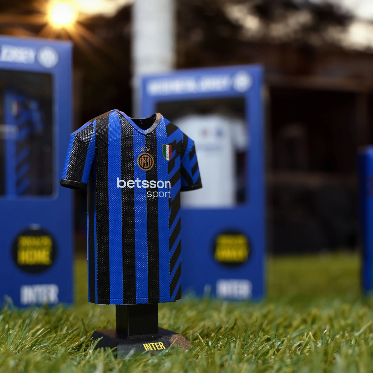 Lifestyle product photograph showcasing an Inter Milan 24/25 jersey collectible standing upright on an outdoor turf surface with scenic background lighting.