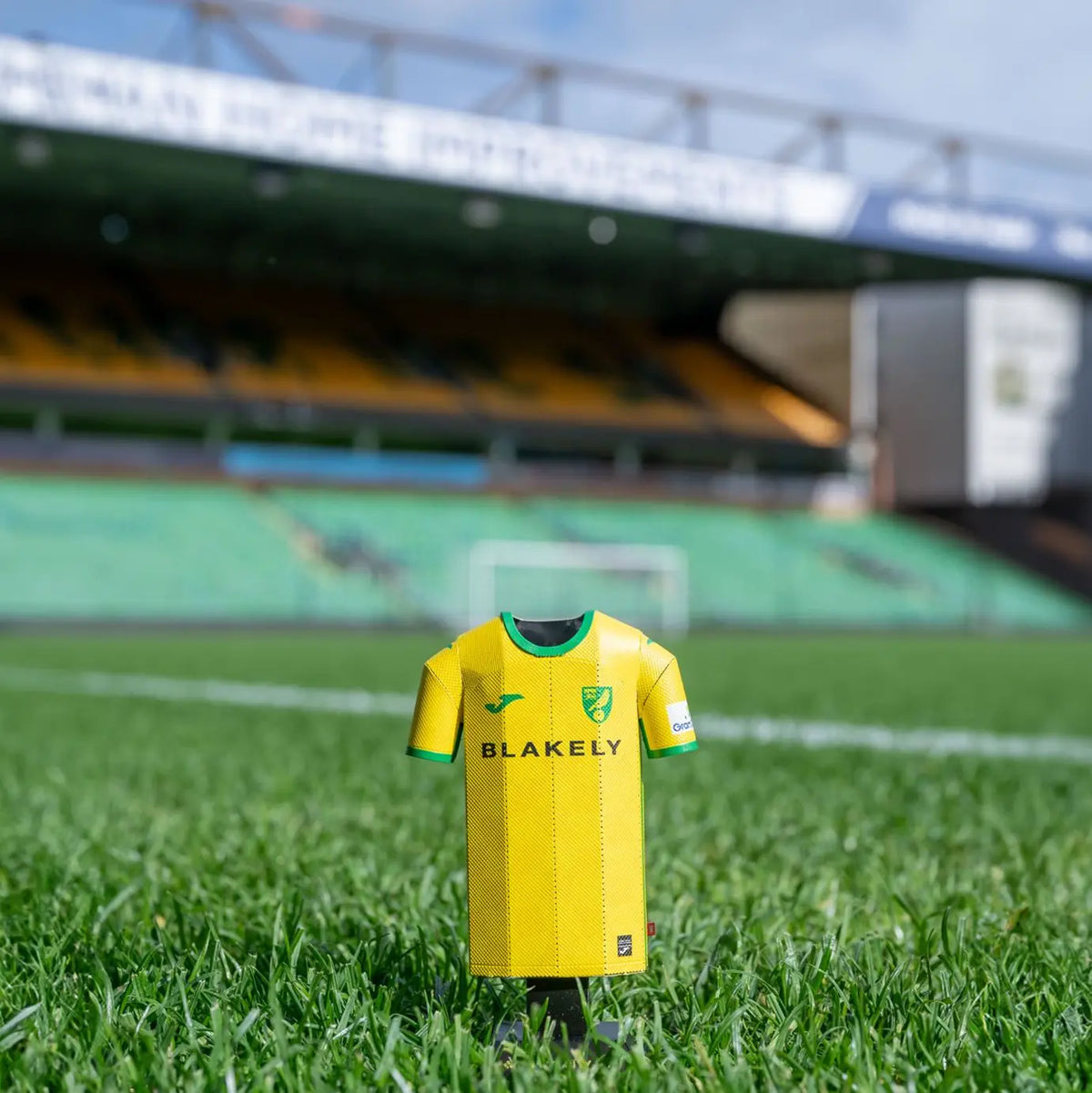 Lifestyle product photograph showcasing a Norwich City FC 24/25 yellow home jersey collectible standing on an outdoor surface near the stadium.
