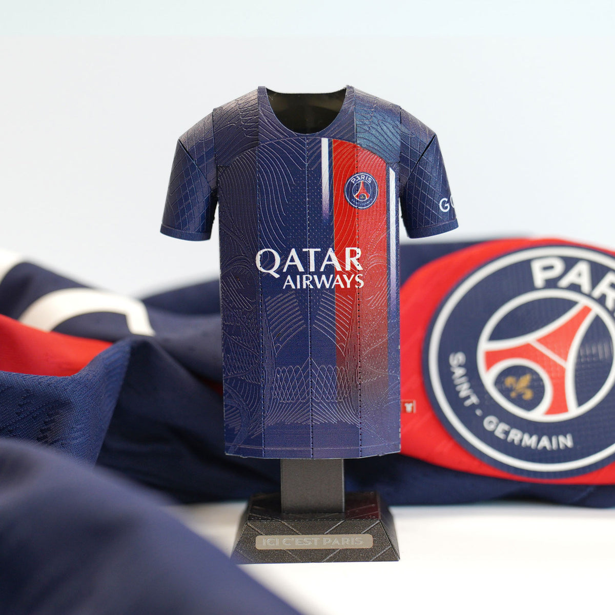 A close-up product photograph showcasing the classic dark blue and red color blocking details of a Paris Saint-Germain 2023/2024 home jersey collectible, displayed next to its branded packaging