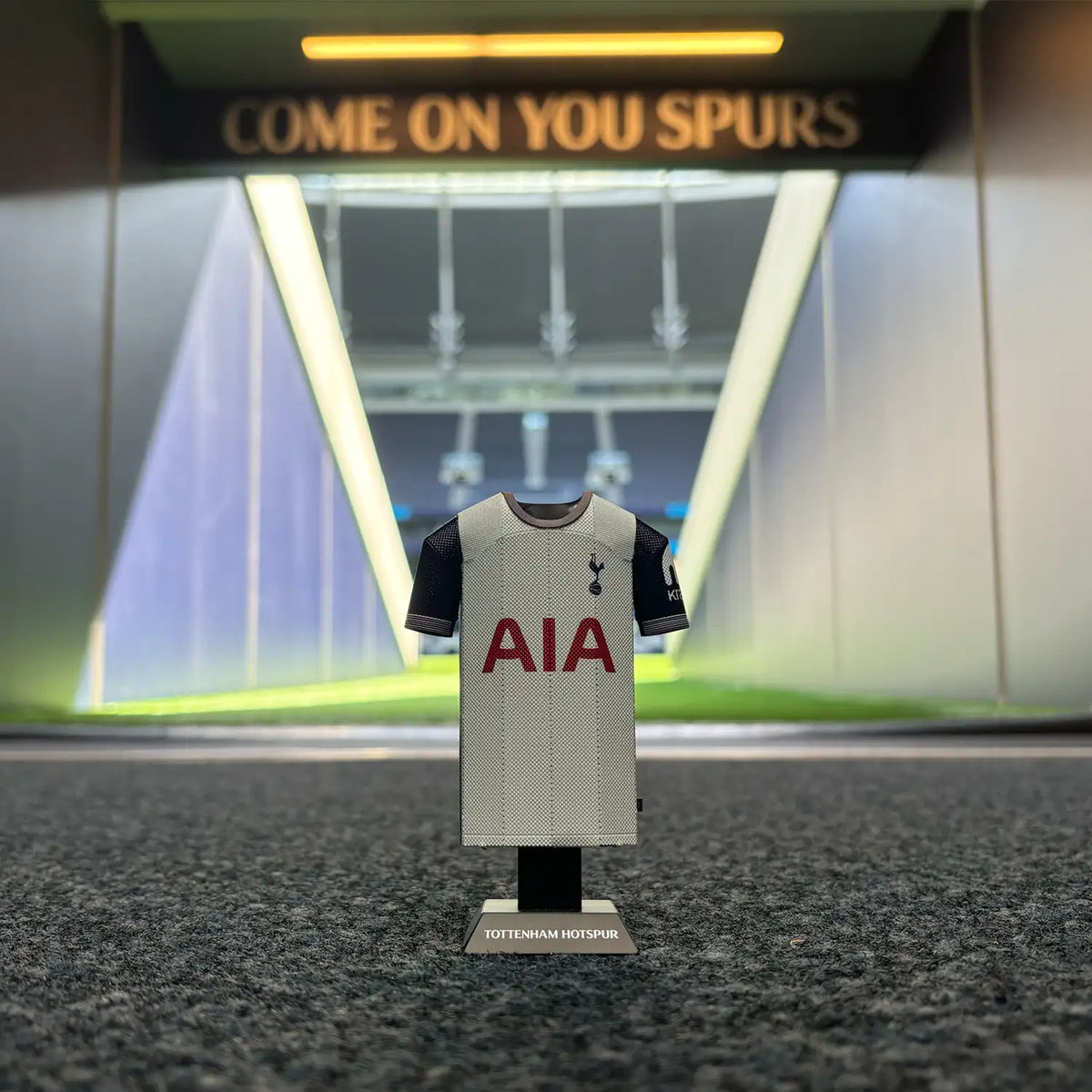 A graphic mockup or lifestyle scene showcasing a Tottenham Hotspur home jersey collectible set against a stylized, illuminated modern stadium tunnel background.