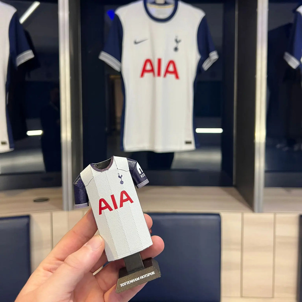 A close-up photograph of a hand holding a premium, officially licensed Tottenham Hotspur miniature product in front of a collectible jersey display background