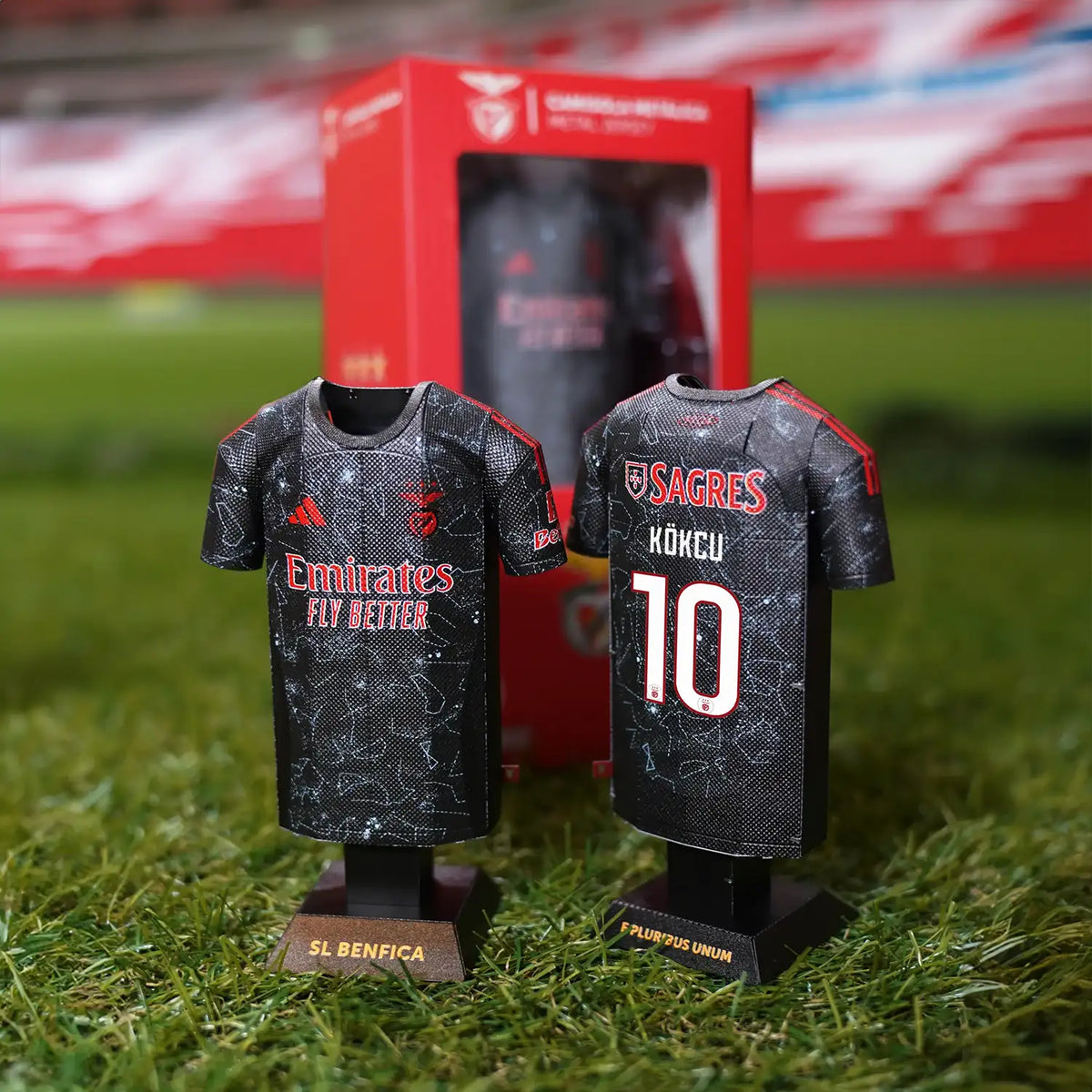 Lifestyle product photograph showcasing a collection of SL Benfica 2024/2025 jersey collectibles standing next to their branded display packaging on outdoor turf.