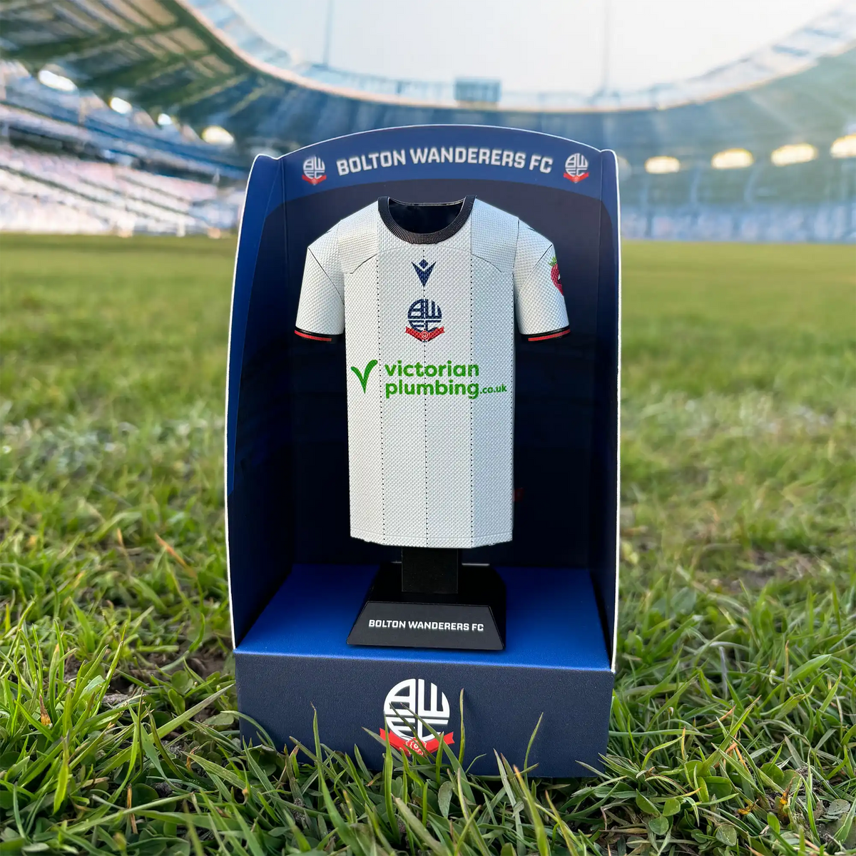 Square lifestyle product photograph showcasing a Bolton Wanderers FC jersey collectible inside its premium display case, set against a stadium background backdrop.