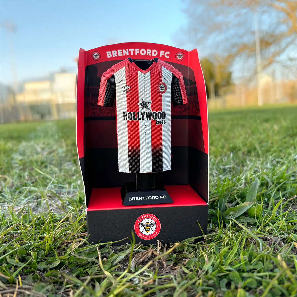 Square lifestyle product photograph showcasing a Brentford FC premium red and white themed display box standing outdoors.