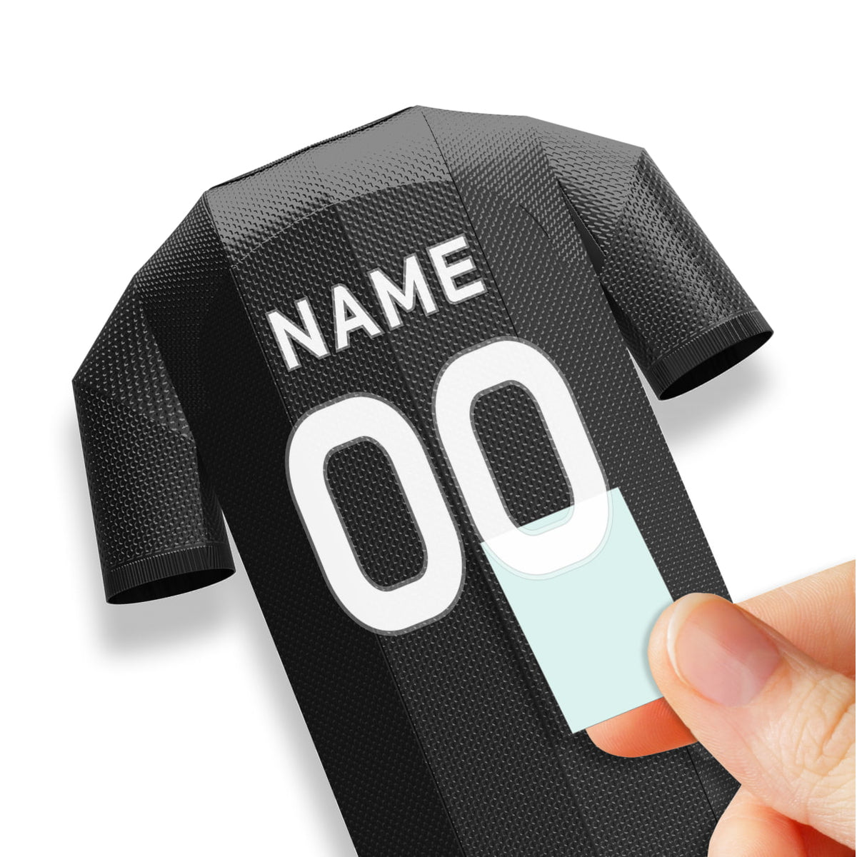 A close-up demonstration photograph showing a hand carefully applying a custom white number decal onto the back of a black miniature jersey model