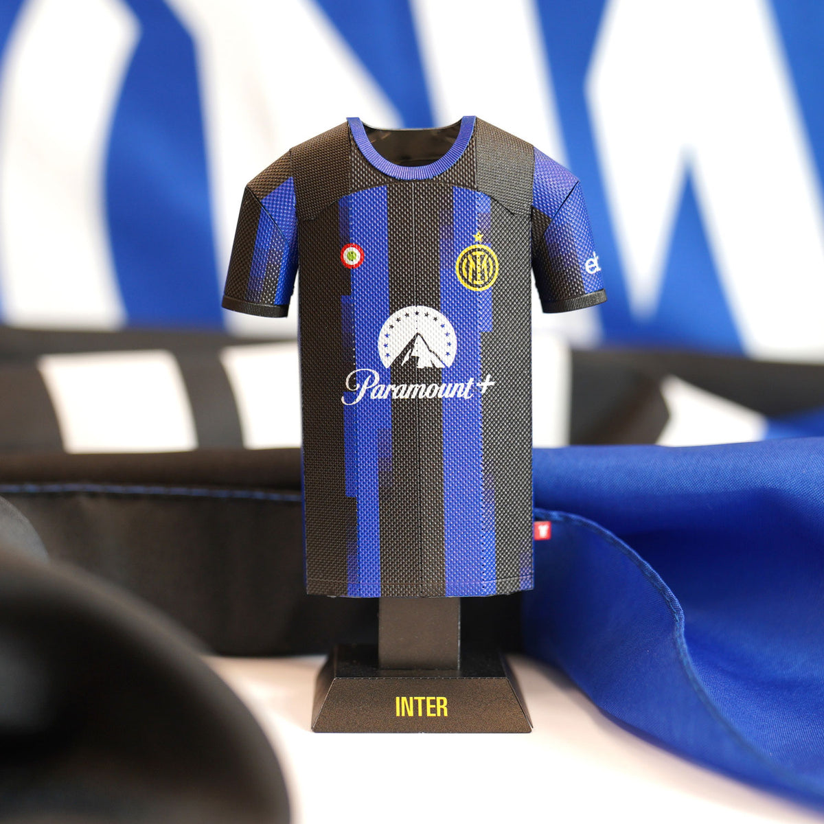 A close-up product photograph showcasing the detailed texture and iconic blue and black vertical stripes of an Inter Milan 2023/2024 home jersey collectible, presented alongside its branded presentation box