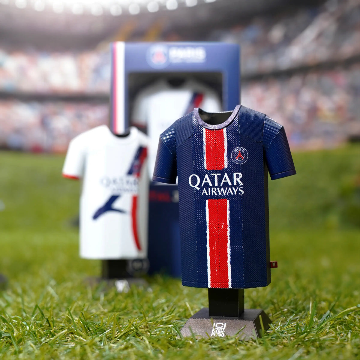 Square lifestyle product photograph showcasing both the home and away Paris Saint-Germain 2024/2025 jersey collectibles standing together on outdoor turf.