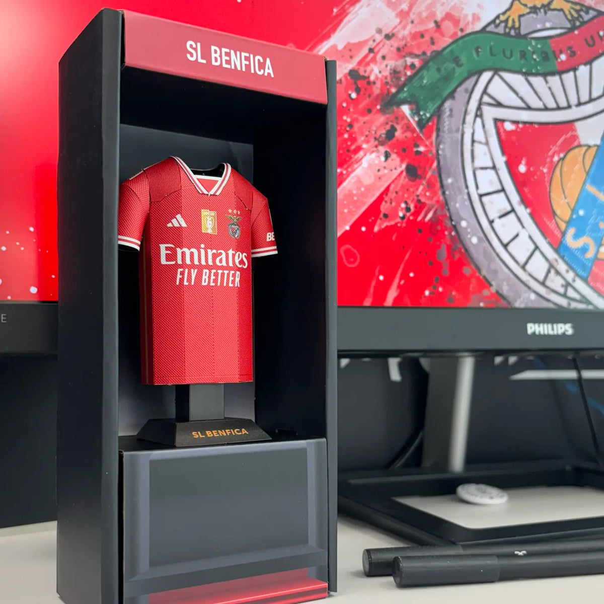 Lifestyle product photograph showcasing an SL Benfica collectible jersey from the 2023/2024 season displayed neatly on a desk surface alongside its presentation box.