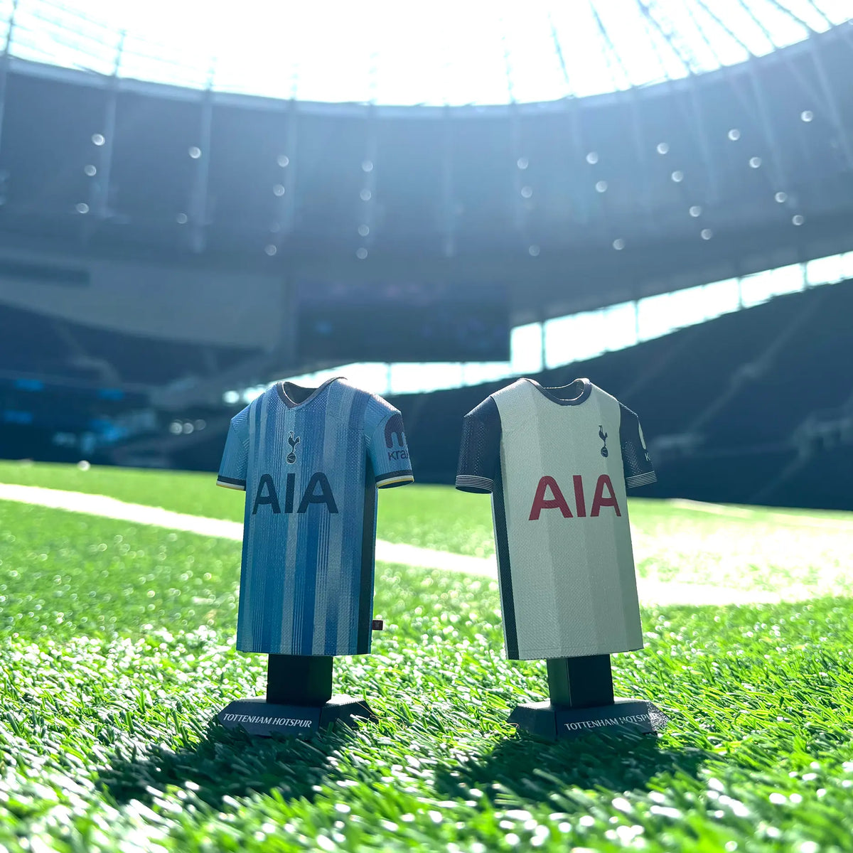 An edited product photograph showing Tottenham Hotspur 3D metal jersey collectibles displayed on outdoor grass overlooking a stadium pitch