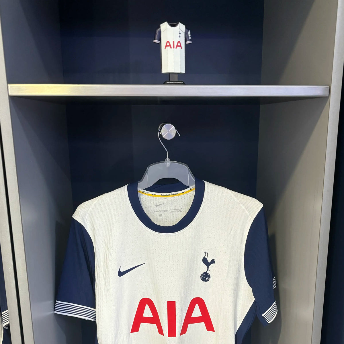 A product presentation graphic showcasing a Tottenham Hotspur white home jersey collectible displayed hanging inside a miniature dark gray stadium locker room stall